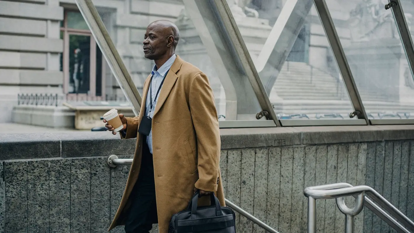 Un salarié en manteau, café et sacoche à la main, sort du métro dans un quartier d'affaires : en Europe, l'exposition à l'IA se concentre dans les emplois de bureau des grandes villes.