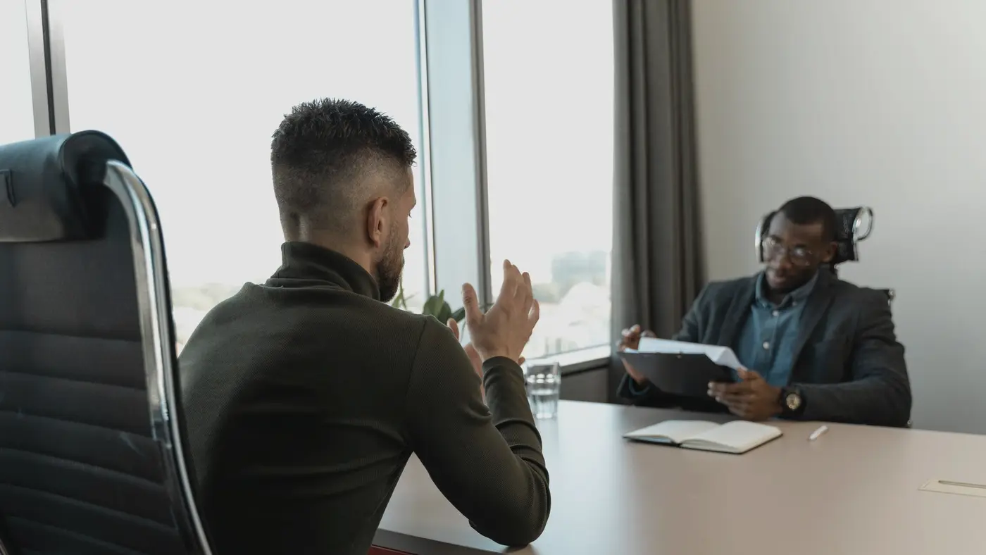 Un entretien d'embauche dans un bureau, un candidat de dos face à un recruteur qui lit un dossier : l'effet de l'IA sur l'emploi se lit d'abord dans les embauches.