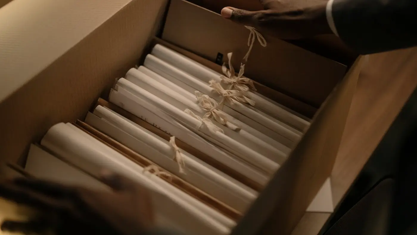 Des mains ouvrent une boîte d’archives remplie de documents roulés et noués : vingt ans de témoignages ne se reconstituent pas.